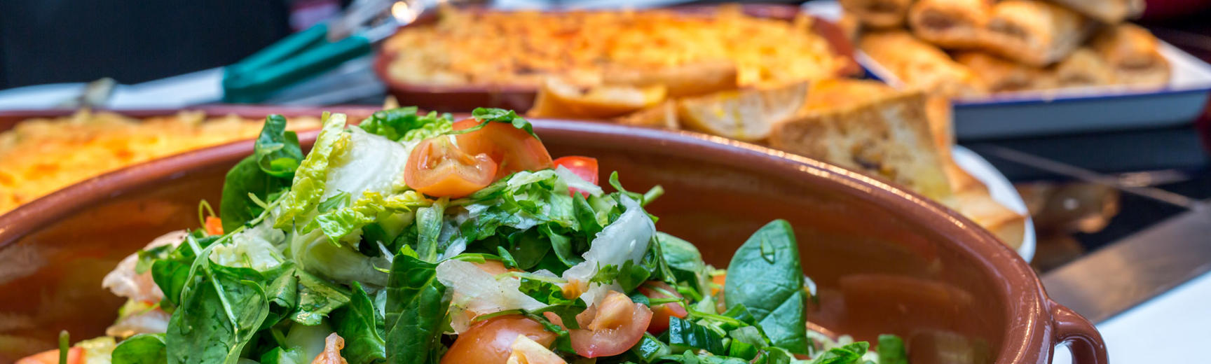 Close-up photo of a bowl of green salad with various hot food in background