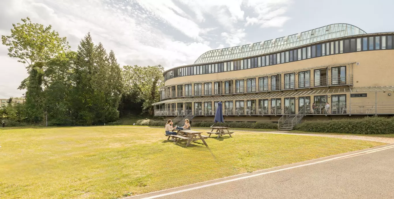 Exterior view of University Club with people sat a picnic bench on lawn eating